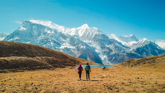 A couple walking towards the Ice lake, as part of the Annapurna Circuit Trek, Himalayas, Nepal. Annapurna chain in the back, covered with snow. Clear weather, dry grass, snowy peaks. High altitude